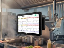 A Kayana wall-mounted touchscreen in a commercial kitchen displays a grid of color-coded pending order tickets.
