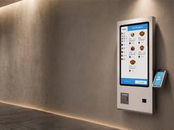 Wall-mounted white self-service kiosk displaying a digital food menu with item photos, prices, and checkout controls.