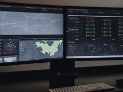 Two desktop monitors display a dark operations dashboard with maps, route-like diagrams, colored status indicators, and tabular lists.