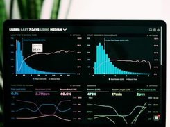 A monitor displays a dark analytics dashboard with histogram charts, trend lines, and metrics about users’ median usage over the last seven days.