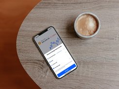 A phone on a table beside a coffee cup displays a Bitcoin chart and a percent-change alert configuration panel.